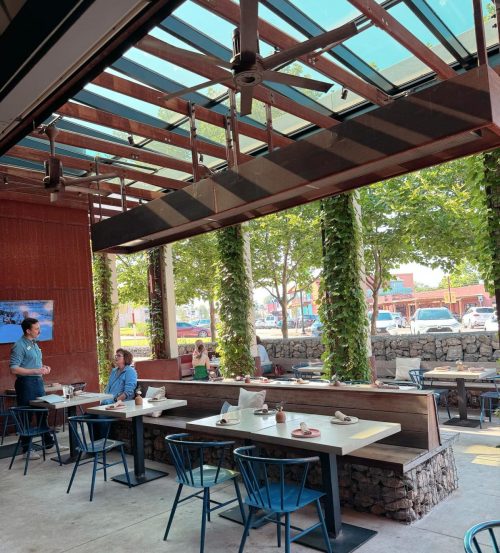 A modern restaurant interior with large windows, greenery, and diners at tables, creating a bright and inviting atmosphere.