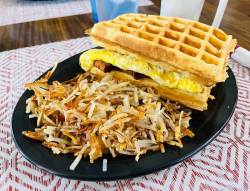 A plate with a waffle sandwich filled with egg and bacon, accompanied by a side of crispy hash browns.