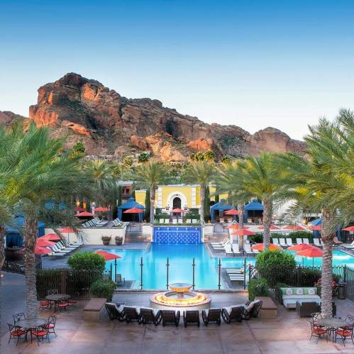 A resort pool surrounded by palm trees, colorful buildings, and mountains in the background under a clear blue sky.