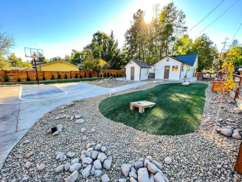 A landscaped backyard featuring a basketball court, a grassy area, and a small shed surrounded by rocks and trees.