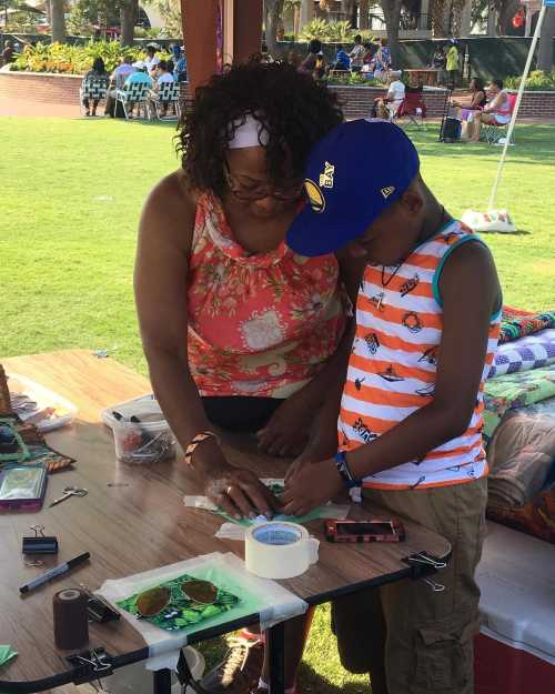 A woman and a boy work together at a table, crafting with materials in a sunny outdoor setting.