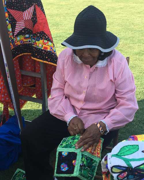 An elderly woman in a black hat sits outdoors, sewing colorful quilt patches with vibrant fabrics around her.