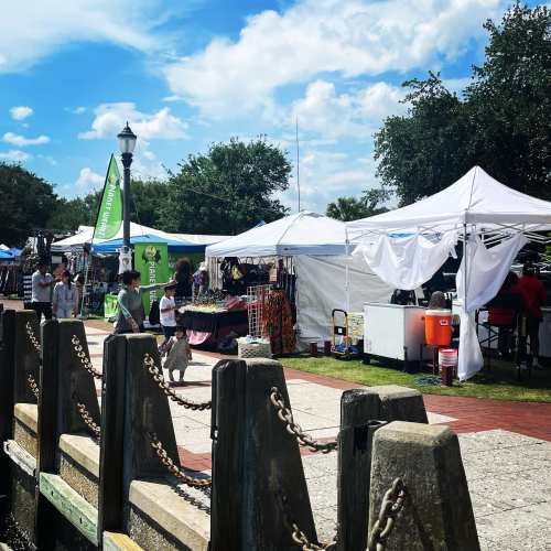 A vibrant outdoor market with tents, vendors, and visitors enjoying a sunny day by the waterfront.