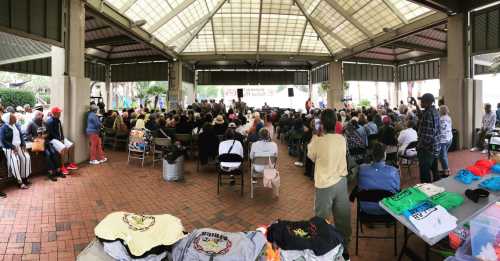 A large crowd gathers under a pavilion for an event, with colorful shirts displayed in the foreground.