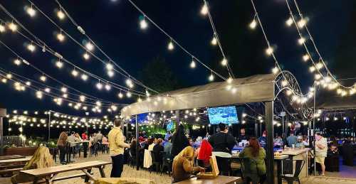 A lively outdoor dining area at night, illuminated by string lights, with people enjoying food and watching a screen.