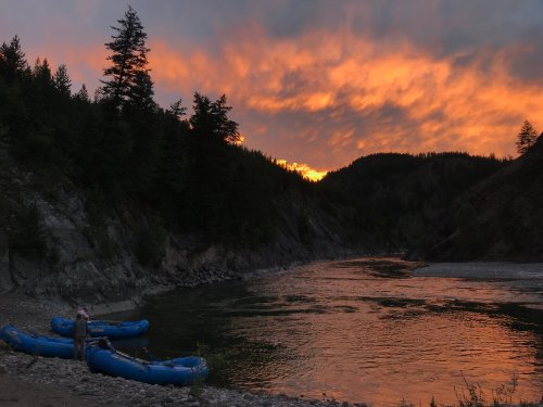 A serene river scene at sunset, with rafts on the shore and vibrant orange clouds above a forested landscape.