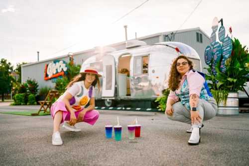 Two women pose in front of a silver trailer, crouching beside colorful drinks on the ground, surrounded by greenery.