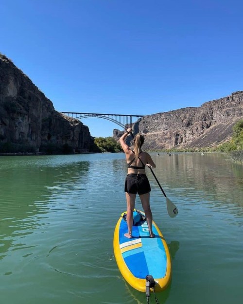 A person paddleboarding on a calm river with a bridge and rocky cliffs in the background under a clear blue sky.