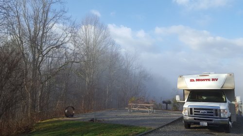 A parked RV in a misty forest setting, with a picnic table and fire pit nearby under a cloudy sky.