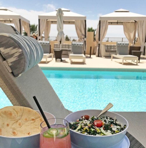 A refreshing poolside scene featuring a colorful drink, bowls of salad, and flatbread, with cabanas in the background.