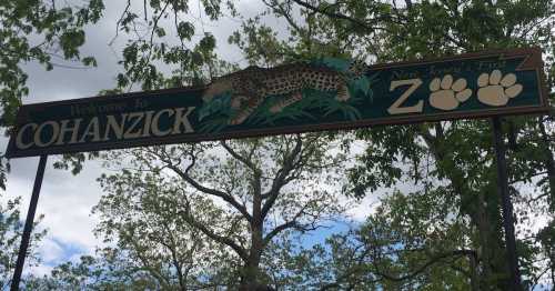 Sign welcoming visitors to Cohanzick Zoo, featuring a leopard and paw prints, surrounded by trees and a cloudy sky.