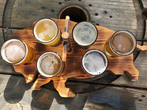 A wooden tray shaped like a pig holds six glasses of different beers on a rustic table.