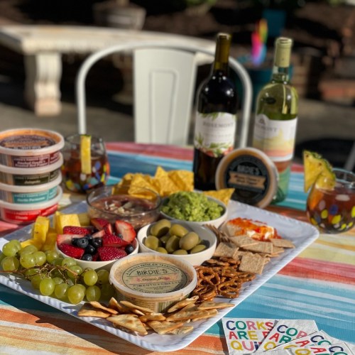 A colorful spread of snacks, dips, fruits, and wine bottles on a table, with drinks and decorative coasters.