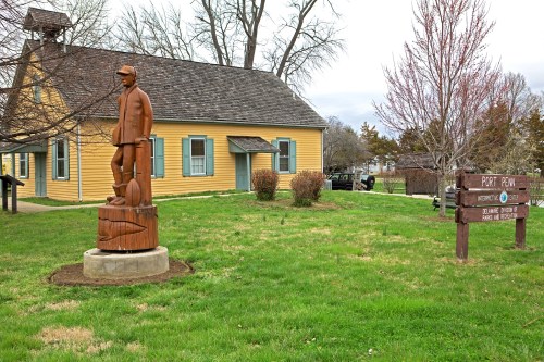A wooden statue of a man stands on a pedestal in front of a yellow historic house and a sign for Port Penn.