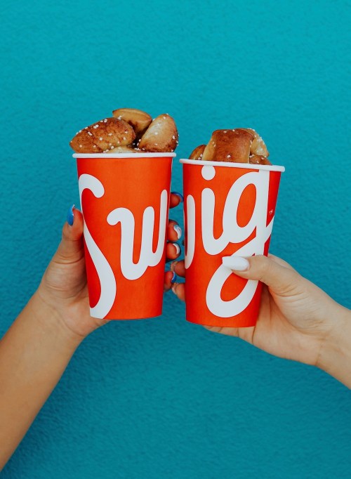 Two hands hold bright red cups with the word "Swig," filled with pretzel bites, against a turquoise background.
