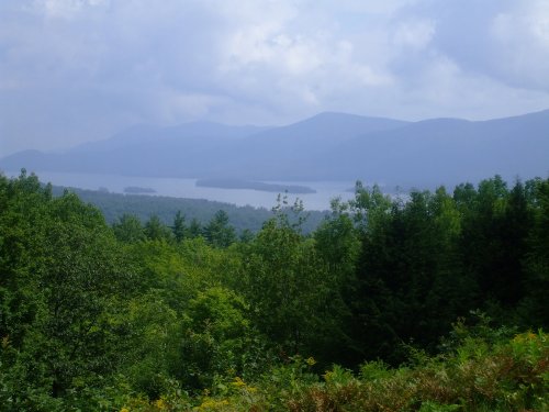 A scenic view of mountains and a lake, surrounded by lush green trees under a cloudy sky.