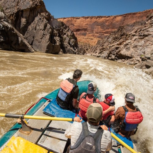 A group of people in a raft navigating a river through a rocky canyon under a clear blue sky.