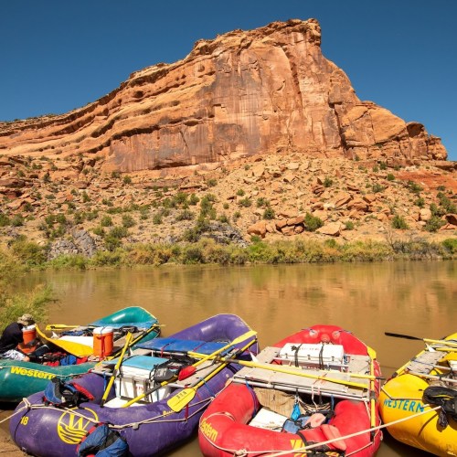 Colorful rafts are docked by a river, with a rocky cliff and clear blue sky in the background.
