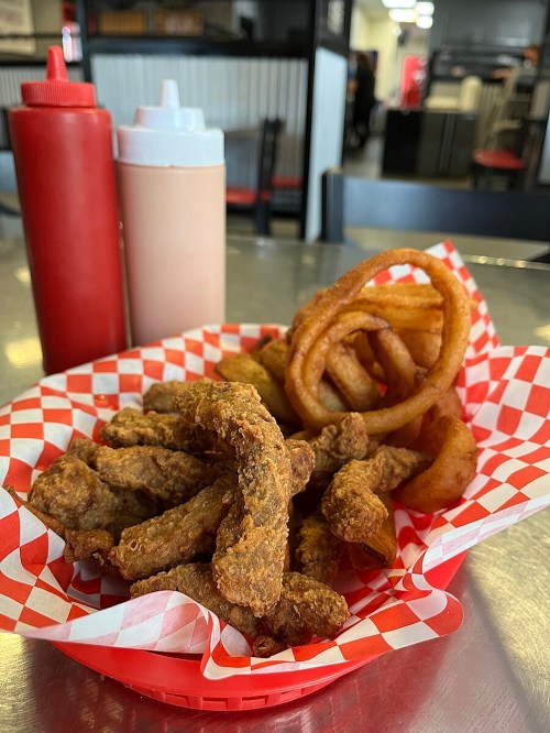 A basket of crispy fried food, including onion rings and strips, with ketchup and sauce bottles in the background.