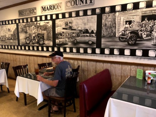 A diner with a man sitting alone at a table, surrounded by a mural depicting historic Marion County scenes.