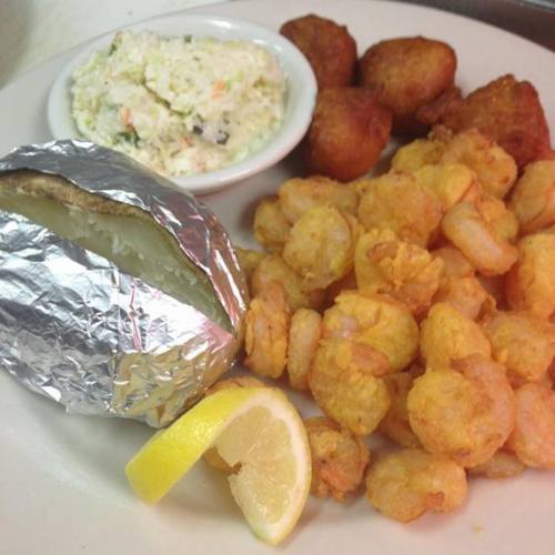 A plate of fried shrimp, a baked potato wrapped in foil, coleslaw, and hushpuppies, garnished with a lemon wedge.