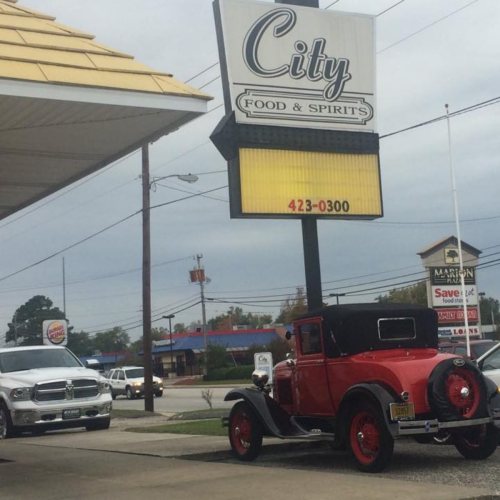A vintage red car parked near a sign for City Food & Spirits, with a cloudy sky in the background.
