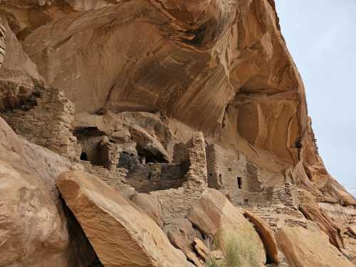 Ancient stone ruins nestled under a large rock overhang, surrounded by rugged terrain and sparse vegetation.