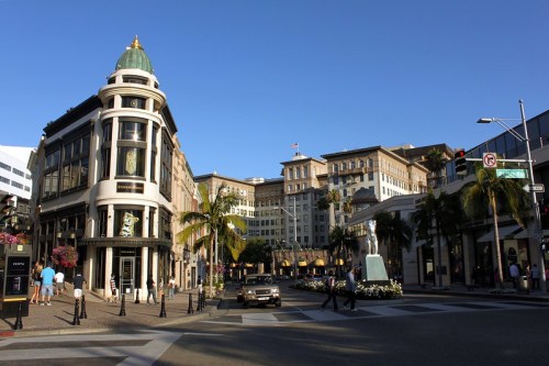 A sunny street view of a luxury shopping area with palm trees, historic buildings, and pedestrians.