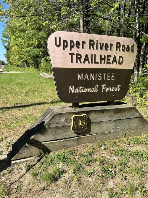 Sign for Upper River Road Trailhead in Manistee National Forest, surrounded by greenery and blue skies.