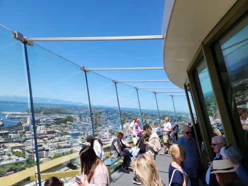 A panoramic view from a glass observation deck with people enjoying the scenery on a sunny day.
