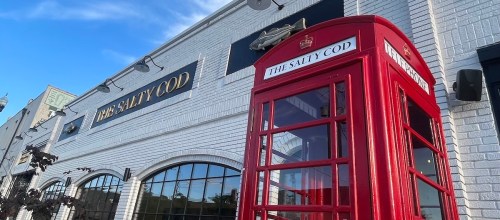 A red telephone booth stands in front of a white building with "The Salty Cod" sign. Clear blue sky above.