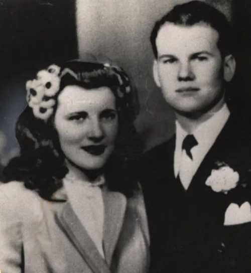 A vintage black-and-white photo of a couple, dressed formally, smiling together. The woman has flowers in her hair.