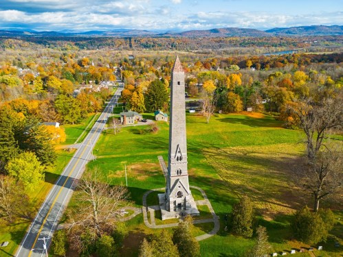 Aerial view of a tall stone tower surrounded by colorful autumn trees and a winding road in a rural landscape.