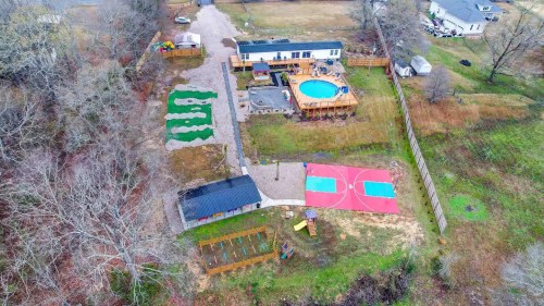 Aerial view of a property featuring a pool, basketball court, garden beds, and a house surrounded by trees.