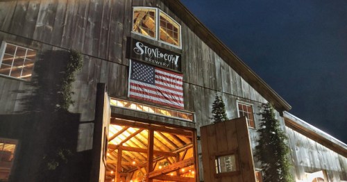 A rustic brewery building at night, featuring an American flag and warm lights glowing from inside.