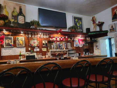 A cozy bar with a wooden counter, red stools, festive decorations, and vintage posters on the walls.