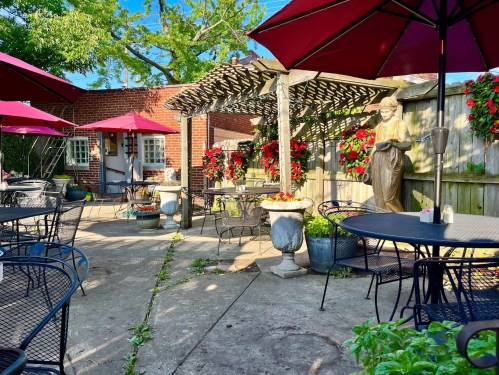 A charming outdoor patio with red umbrellas, flower pots, and a statue, surrounded by greenery and brick walls.