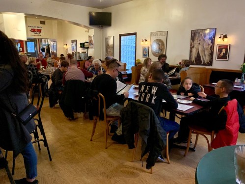 A busy restaurant interior with diners at tables, some looking at menus, and a warm, inviting atmosphere.