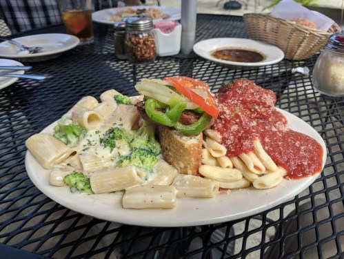 A plate of pasta with broccoli, marinara sauce, and a meatball, served on a black outdoor table.