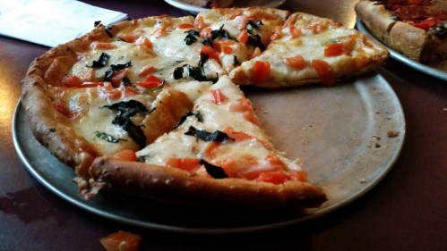 A pizza on a metal tray, topped with melted cheese, tomatoes, and basil, with a slice removed.