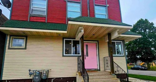 A modern two-story building with red and beige siding, a pink door, and a green roof. Steps lead to the entrance.
