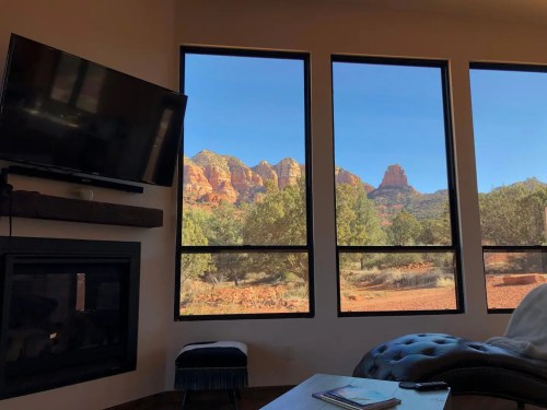 View of red rock formations through large windows, with a cozy interior featuring a TV and a fireplace.