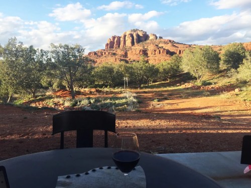 A scenic view of red rock formations and greenery, with a glass of red wine on a table in the foreground.