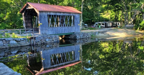 A wooden covered bridge reflects in calm water, surrounded by lush greenery and a peaceful campsite.