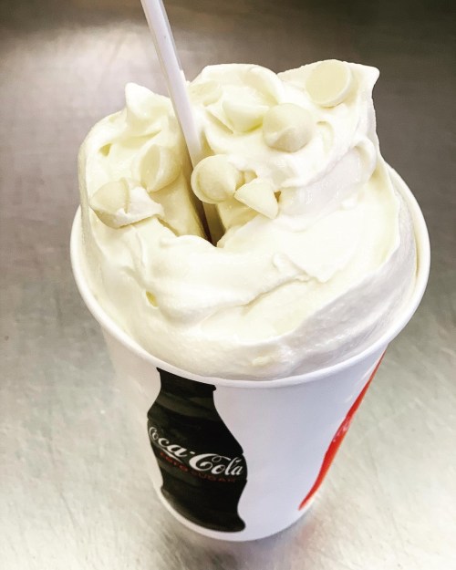 A cup of creamy white soft serve ice cream topped with white chocolate chips and a straw.