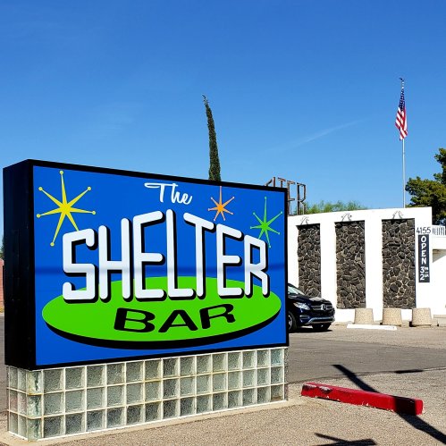 A colorful sign for "The Shelter Bar" with a blue background, green accents, and a nearby American flag.