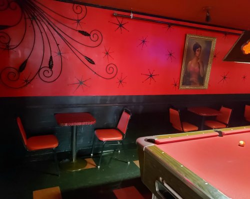 A retro-style bar with red walls, a vintage painting, and a pool table, featuring black and red seating.