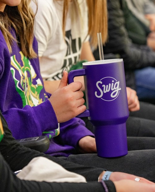 A person holds a large purple Swig cup with a straw, sitting among others in a casual setting.