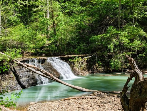 A serene waterfall cascades into a turquoise pool, surrounded by lush green trees and rocky terrain.