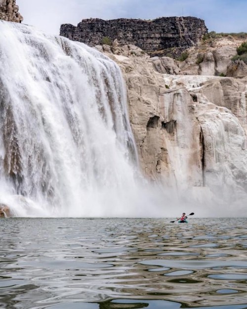 A kayaker paddles near a large waterfall, surrounded by rocky cliffs and calm water.